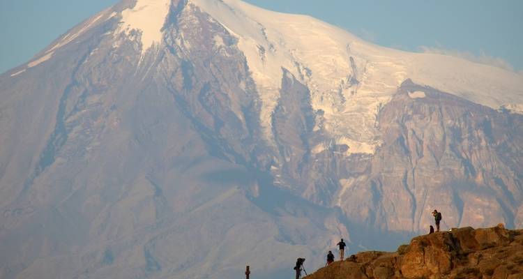 A towering snow-capped mountain with rocky terrain and hikers in the foreground.