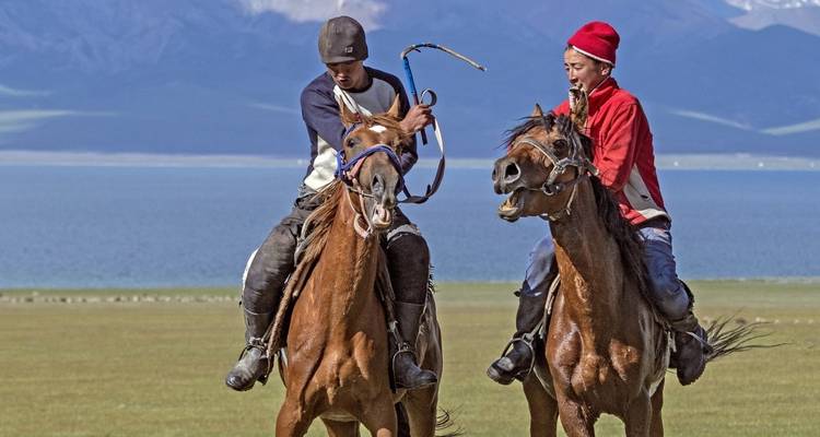 Two people on horseback near a lake with mountain backdrop.