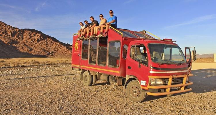 Group sitting on top of a red truck in a desert landscape.