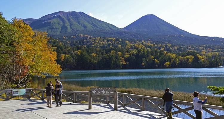 People standing by a lake with a view of mountains and autumn trees.