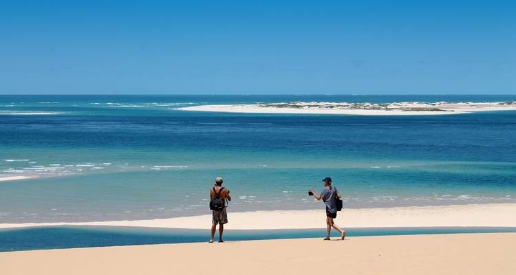 Two people walking on a sandy beach with a clear blue ocean and a sandbar in the distance.
