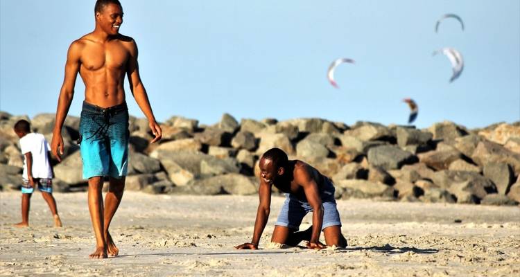People on a sandy beach with rocks and kites in the background.