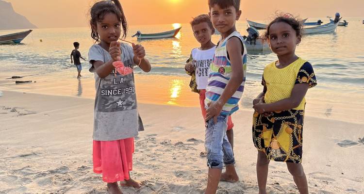 Children playing on a beach at sunset with boats in the background.