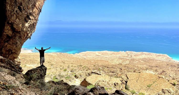 Person standing with arms raised on a cliff overlooking the ocean.