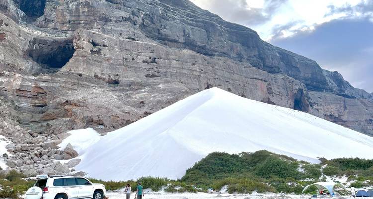 White sand dune with a car and small group of people at the base of a large rock formation.