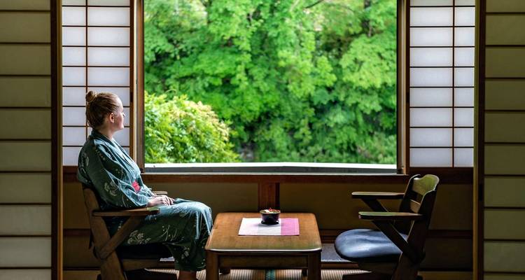 Person in traditioneller Kleidung, die üppiges Grün von einem Fenster aus betrachtet.