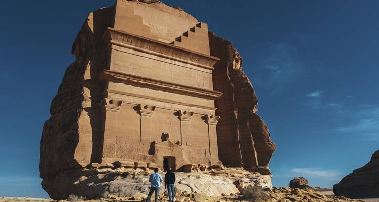 Large rock-hewn tomb with two visitors standing in front.
