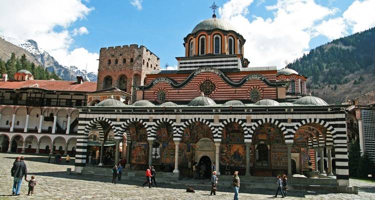 Rila Monastery with people walking around, surrounded by mountain scenery.