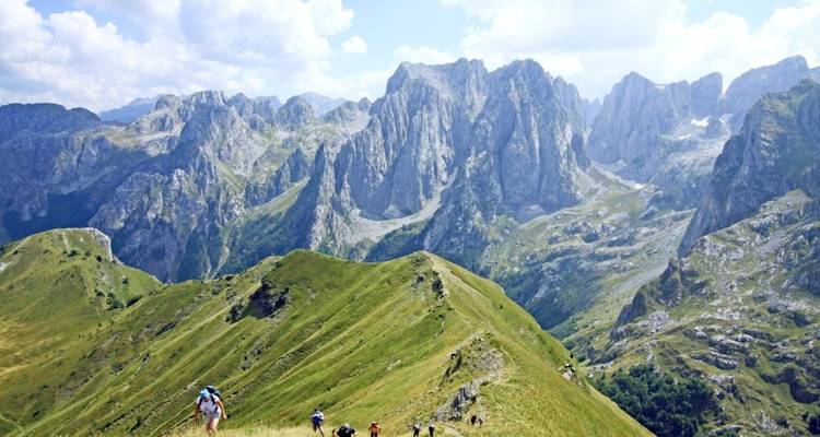 A group of hikers walking on a grassy ridge with towering mountains in the background.