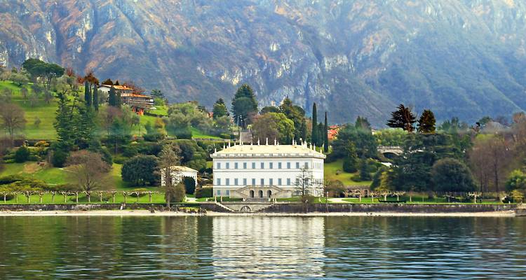 Eine stattliche weiße Villa inmitten sanft geschwungener grüner Gärten unter schroffen Bergen am Comer See.