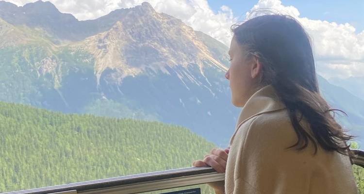 Junge Frau in eine Decke gehüllt blickt von einem Balkon über bewaldete Berge und entfernte Gipfel.