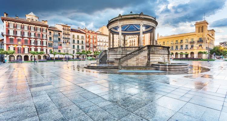 Een stadsplein met een centrale tuinkoepel omringd door kleurrijke gebouwen, weerkaatsend op natte trottoirs.