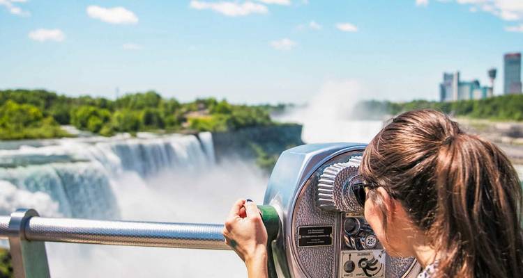 Un visitante mira a través de un visor las brumosas cascadas de las Cataratas del Niágara con el horizonte de la ciudad al fondo.