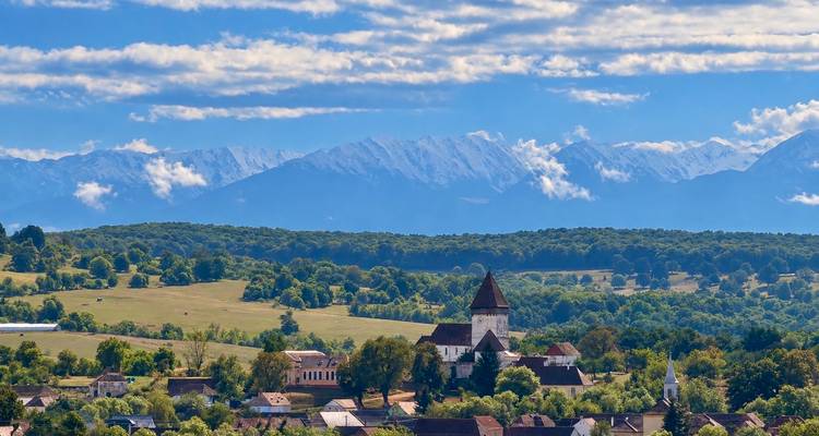 Village roumain pittoresque avec une église fortifiée situé dans des champs ondulants face aux montagnes de Făgăraș.
