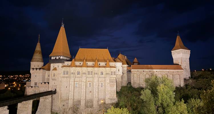 Le château de Corvin illuminé se dresse majestueusement la nuit, ses tours brillant au-dessus des arbres environnants.