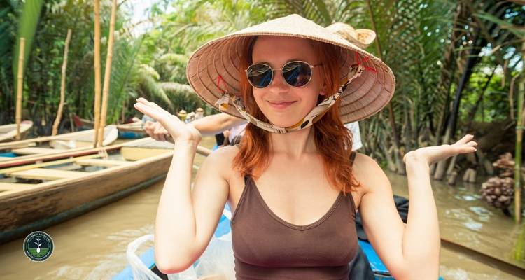 Smiling woman wearing a hat, sitting in a boat surrounded by greenery.