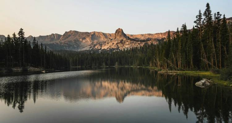 Lac alpin immobile comme un miroir bordé d'arbres à feuilles persistantes denses avec des pics escarpés capturant une lumière chaude.