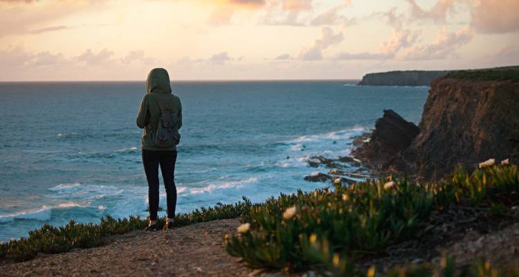 Person standing on a cliff edge overlooking the ocean at sunset.