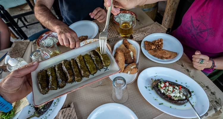 Mesa servida con meze griego incluyendo hojas de parra rellenas y cerveza con comensales alcanzando la comida