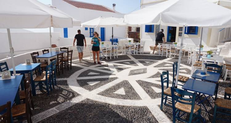 Café de patio abierto con suelo de mosaico de guijarros, sillas azules y paredes encaladas bajo la luz solar brillante
