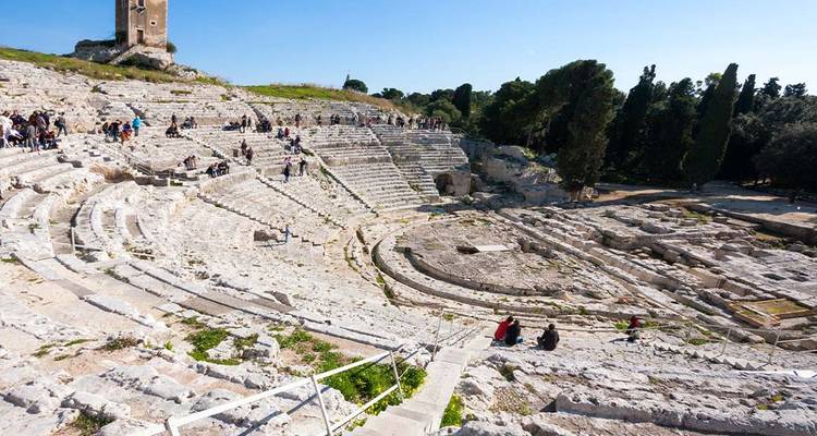 Oud stenen amfitheater uitgehouwen in een heuvel met bezoekers die de getrapte zitplaatsen verkennen onder een heldere blauwe hemel.