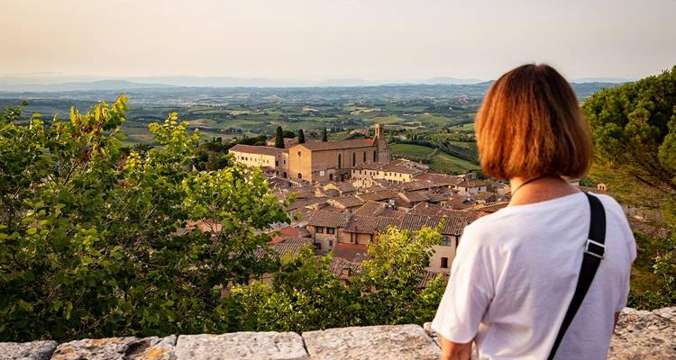 Jonge vrouw die uitkijkt over een Toscaanse stad op een heuveltop met pannendaken en groen landschap in zacht avondlicht.