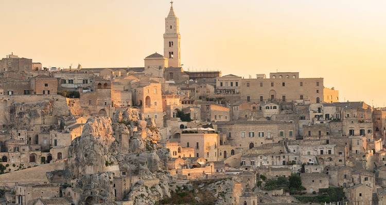Goudverlicht panorama van Matera's stenen grotwoningen en kerktoren die boven ruige kliffen uitrijzen bij schemering.