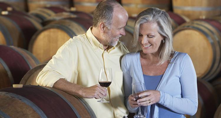 Pareja madura sonriente brindando con copas de vino entre barriles de madera en una bodega