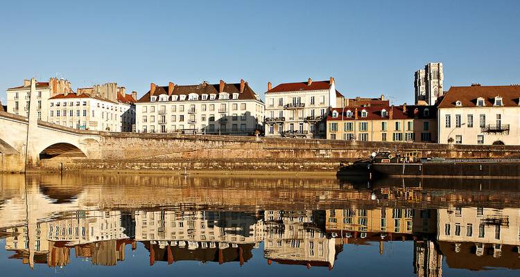 Reihe von Stadthäusern am Flussufer in Burgund, die sich in ruhigem Wasser unter einem klaren blauen Himmel spiegeln.