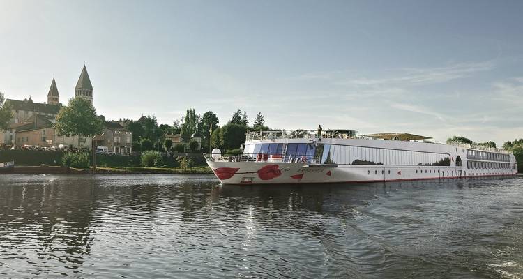 Modernes Flusskreuzfahrtschiff gleitet auf einem ruhigen Fluss an einem malerischen europäischen Dorf mit Kirchtürmen unter einem klaren blauen Himmel vorbei