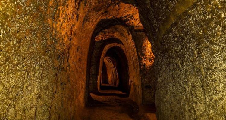 Corridor souterrain sinueux creusé dans la terre, éclairé par des lumières ambre chaud créant de longues ombres.
