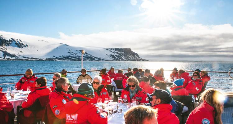 Viajeros con abrigos rojos a juego disfrutando de una comida al aire libre en la cubierta de un barco con acantilados nevados y un sol brillante detrás de ellos.