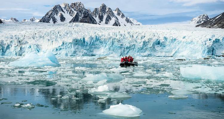 Un pequeño bote inflable con pasajeros en chalecos rojos navega por aguas heladas bajo un glaciar imponente y picos escarpados.