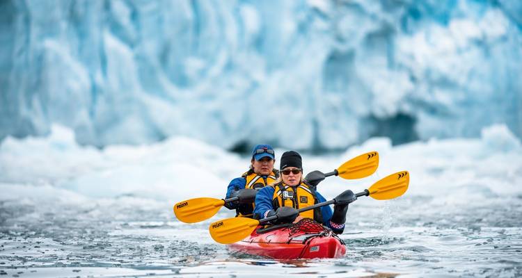 Dos kayakistas reman un kayak rojo a través de aguas árticas heladas con un glaciar azul detrás de ellos.