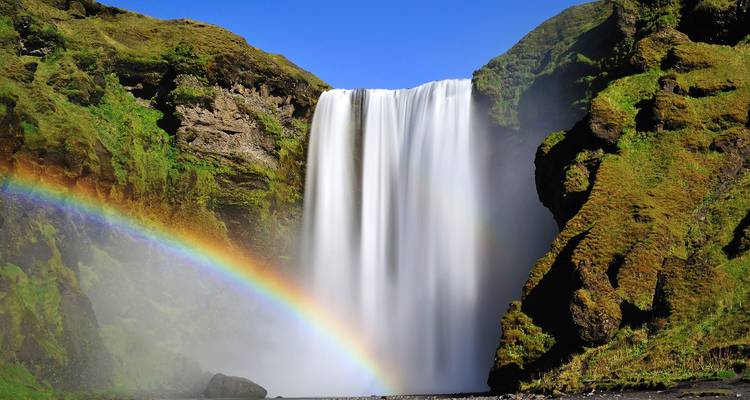 La puissante cascade de Skógafoss avec un arc-en-ciel éclatant contre des falaises mousseuses sous un ciel bleu