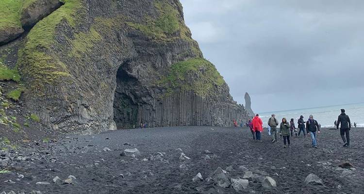 Les visiteurs marchent le long d'une plage de sable noir avec une grotte de basalte et des formations rocheuses en mer sous un ciel gris