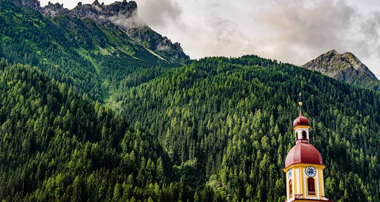 Une tour d'église colorée au dôme rouge se dresse contre un versant montagneux dense de conifères dans le Tyrol autrichien.