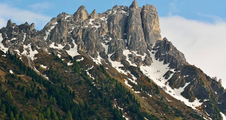 Des pics calcaires déchiquetés tachetés de neige s'élèvent au-dessus de pentes boisées sous un ciel bleu clair.