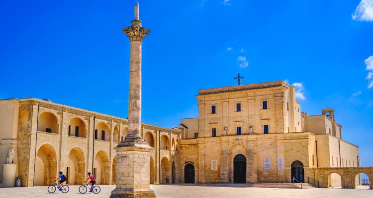 Los ciclistas aran por la enorme plaza frente al santuario de Leuca y la columna bajo un cielo intenso.