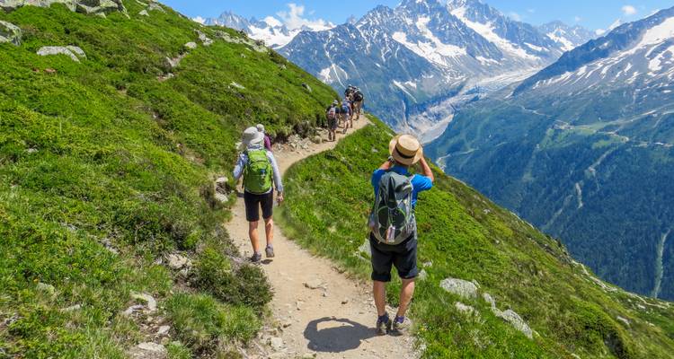 Los excursionistas siguen un sendero montañoso pintoresco a gran altura sobre un profundo valle con los Alpes nevados de fondo.