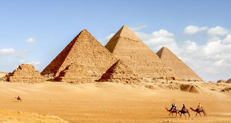Three large pyramids in a desert setting with people riding camels.