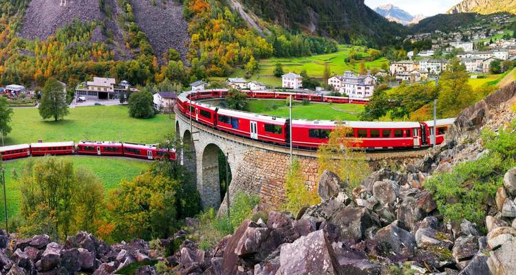 Un tren panorámico de color rojo brillante serpentea a través de un viaducto histórico de piedra entre verdes valles alpinos y follaje otoñal colorido debajo de picos escarpados.