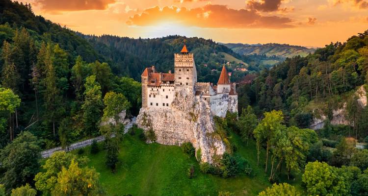 Bran Castle staat op een rotsachtige heuvel omgeven door weelderig bos onder een vurig zonsonderganghemel.