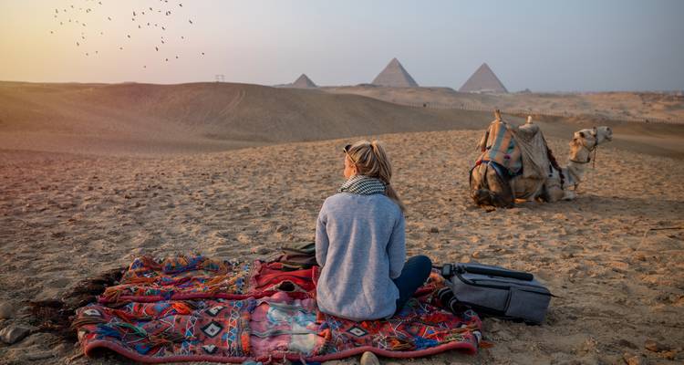 Vrouw zit op een gemusterd kleed in de woestijn met kamelen in de buurt en kijkt naar de piramides tijdens zonsondergang.