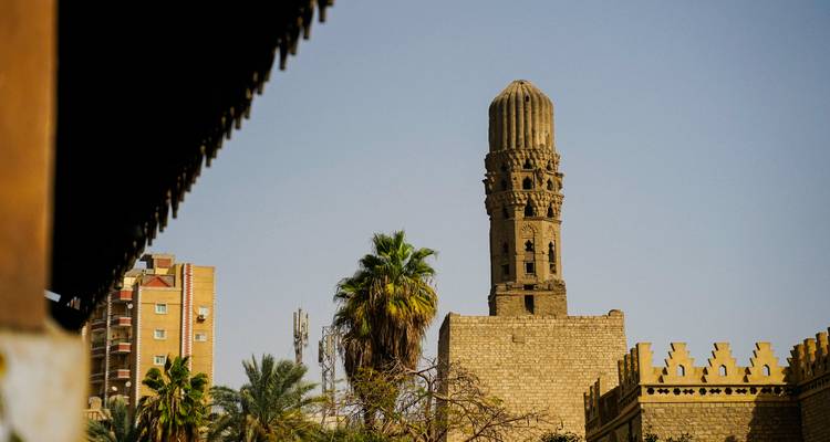 Minarete de piedra que se alza entre palmeras y muros de la ciudad de color arena bajo un cielo brumoso