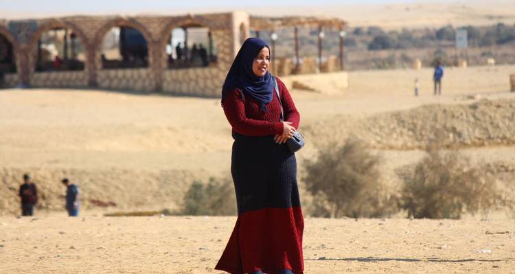 Een vrouw die een hijab en lange jurk draagt staat in een zandige woestijnomgeving met stenen bogen op de achtergrond.