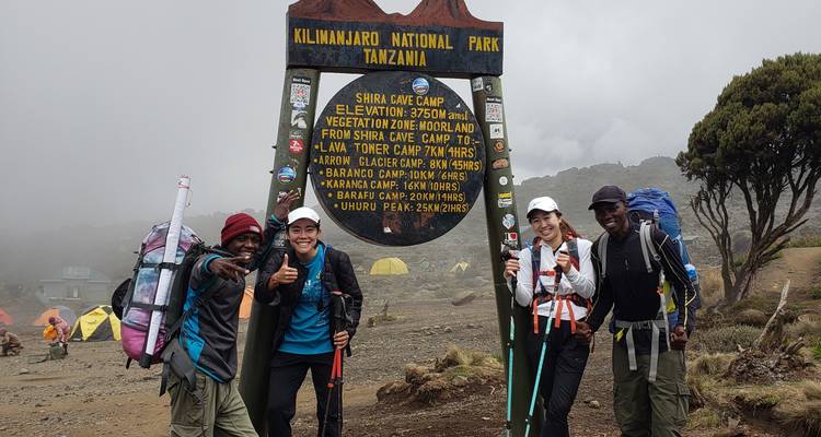 Groep poseert bij een bord voor Shira Camp met tenten op de achtergrond.