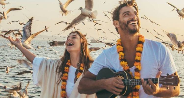 Un couple souriant portant des guirlandes de souci joue de la musique sur un bateau entouré de mouettes volantes au lever du soleil sur le Gange.