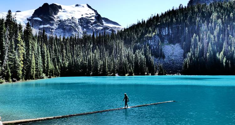 Une personne debout sur une bûche dans un lac turquoise avec des montagnes en arrière-plan.