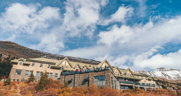 Een berghut met mensen op een balkon en besneeuwde toppen op de achtergrond.